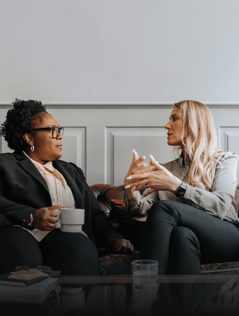 Two women in business attire having a conversation on a leather sofa, one holding a white mug, while the other gestures expressively.