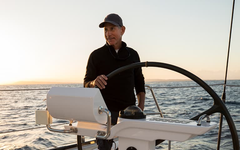 Man in a cap steers a boat at the helm during sunset, with calm ocean waters and a warm glowing horizon stretching behind him.