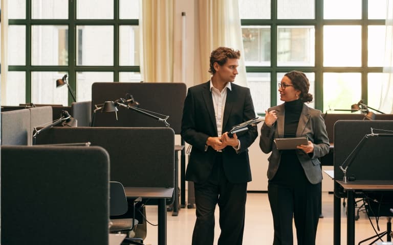 Two business professionals walk through a modern office workspace, conversing while holding tablets, with large green-framed windows and warm natural light.