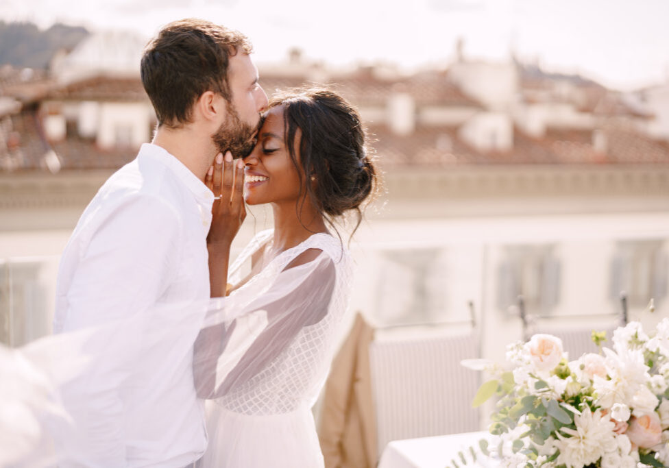 Interracial wedding couple. Destination fine-art wedding in Florence, Italy. Caucasian groom and African-American bride cuddle on the roof, in the sunny sunset light.
