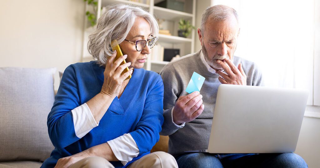Concerned older couple at home on the phone and holding a credit card while reviewing a laptop, representing the risk seniors face from common financial scams and identity theft