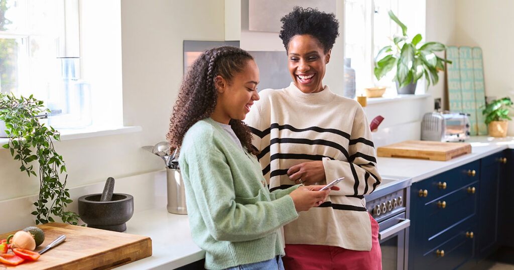 Parent and teenage child standing together in a bright kitchen, talking while looking at a smartphone.