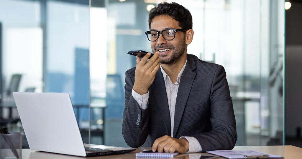 Small business owner speaking into a smartphone at his desk beside a laptop, reflecting the risks of relying on AI for accounting decisions without professional validation