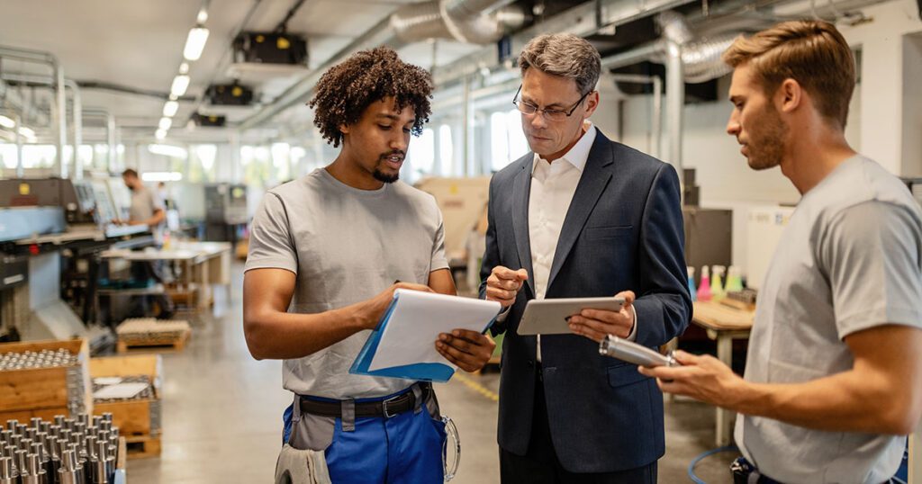Business manager reviewing documents on a tablet with two workers on a manufacturing floor, representing the hidden operational costs of scaling a small business from startup to mid-market