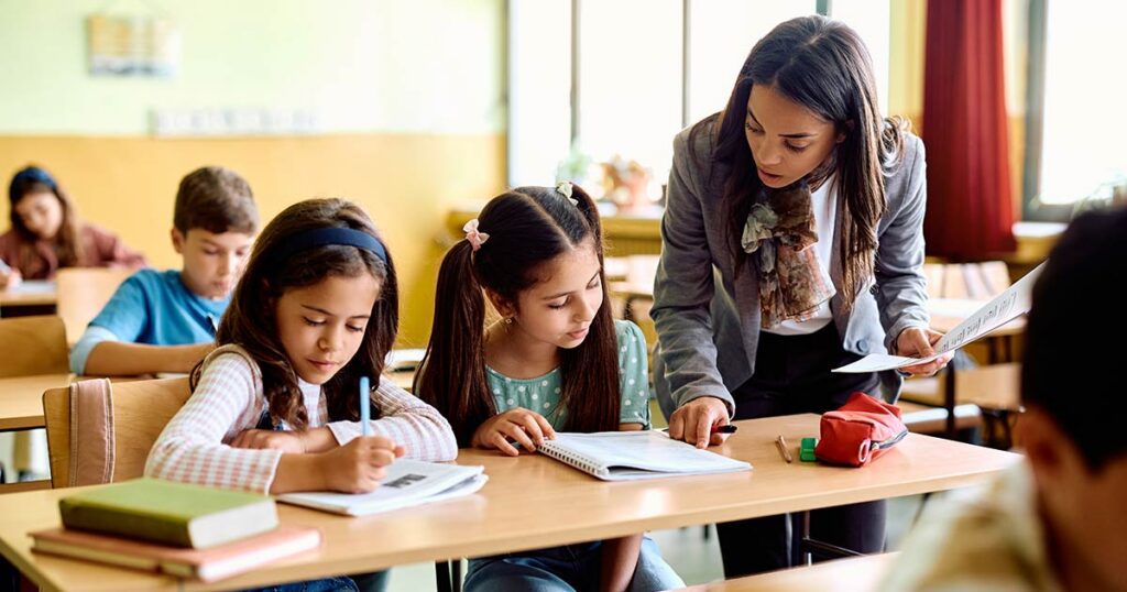Elementary school teacher supporting two young students in class, symbolizing how outsourced school finance services help K‑12 and charter schools focus resources on teaching and learning