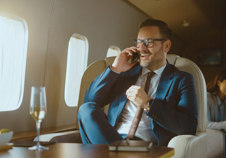 Businessman in a private jet talking on the phone with a glass of champagne on the table.