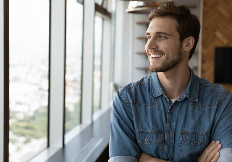 Happy dreaming businessman standing by office or home workplace, looking out of window, thinking of future vision with confidence, smiling at good thoughts, planning investment, enjoying success