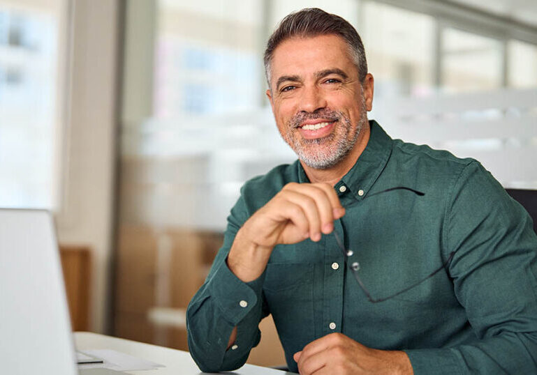 Happy smiling Indian older business man, middle aged professional businessman ceo executive bank manager, mature investor sitting at work desk in office with laptop looking at camera, portrait.