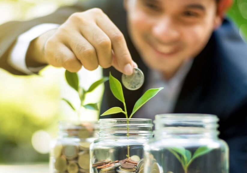 Businessman putting coin into the glass jar with young plant, demonstrating financial growth through saving plans and investment schemes