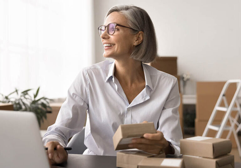 Small business owner sitting at a desk with a laptop, holding product boxes in a workspace filled with packages.