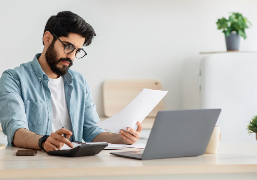 Paying bills, taxes at home online. Young arab man using calculator and laptop computer, calculating taxes or planning budget while sitting at kitchen table at home, copy space